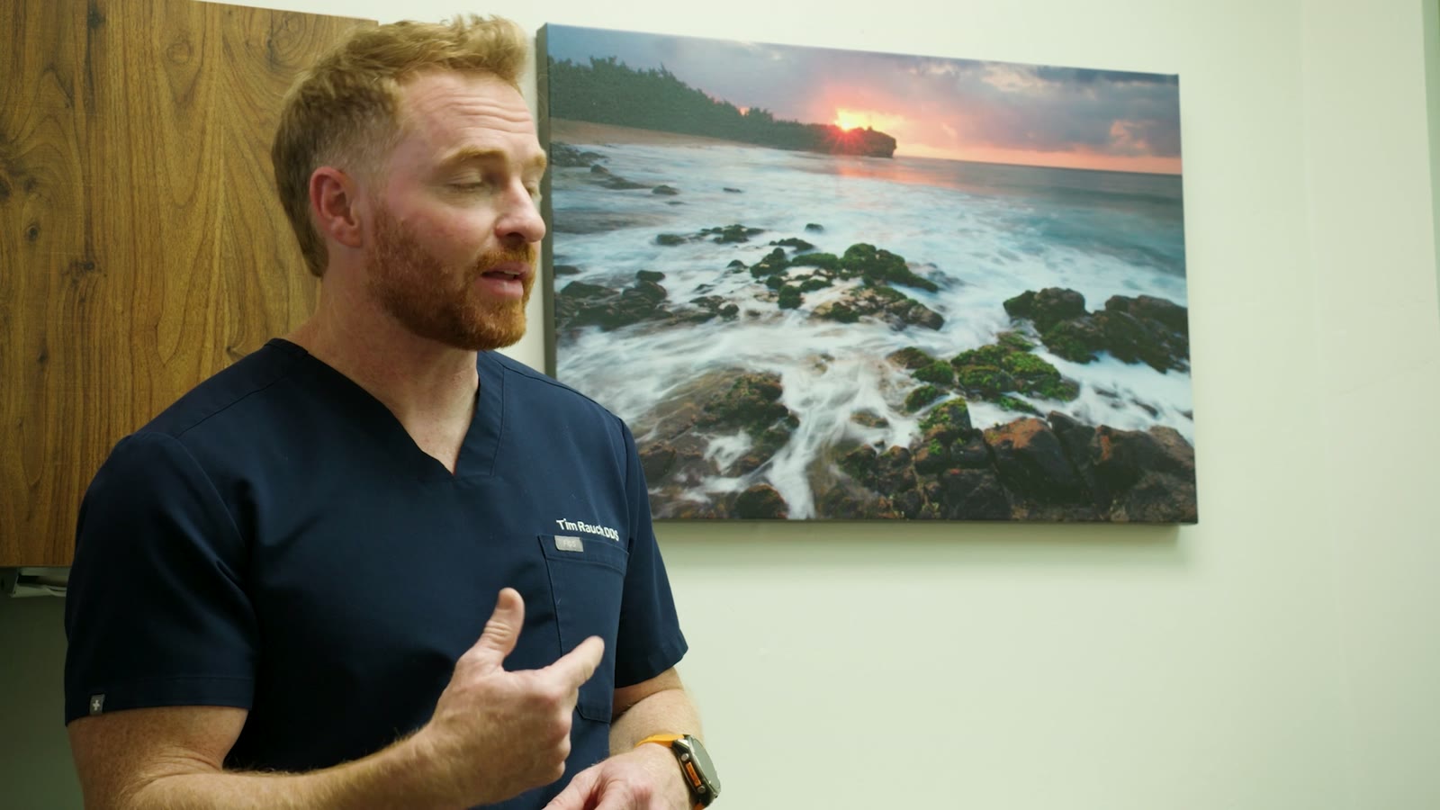 Dr. Tim Rauch in navy scrubs gesturing mid-explanation, coastal art print on the wall